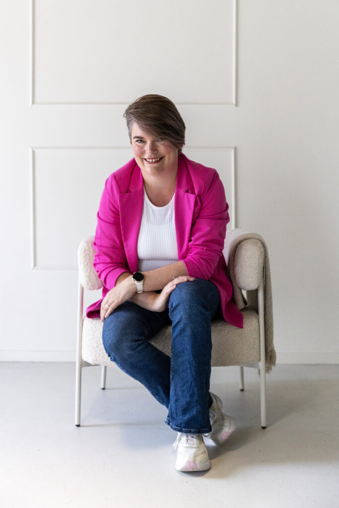 Vrouw in roze jasje zittend op een stoel tegen witte muur